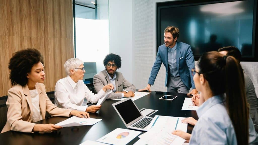 A diverse group of business professionals are gathered around a conference table in an office, discussing documents and charts.
