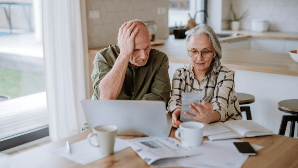 An older man holds his head in his hands as he looks at a laptop while a woman looks at a receipt at a kitchen table.