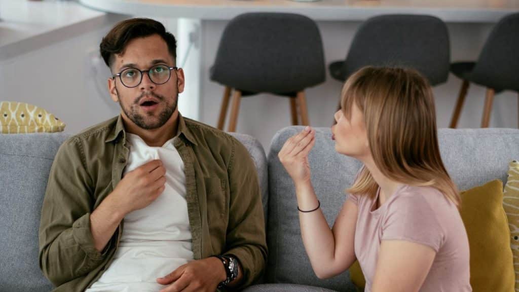 A man with a surprised expression and glasses holds his hand to his chest while a woman gestures and talks to him.