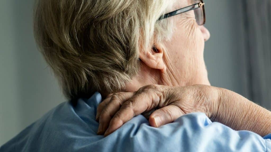A wrinkled hand rests on the shoulder of an older person with gray hair.