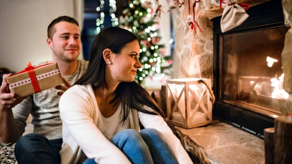 A man is sitting behind a woman, holding a gift as she looks towards a fireplace.