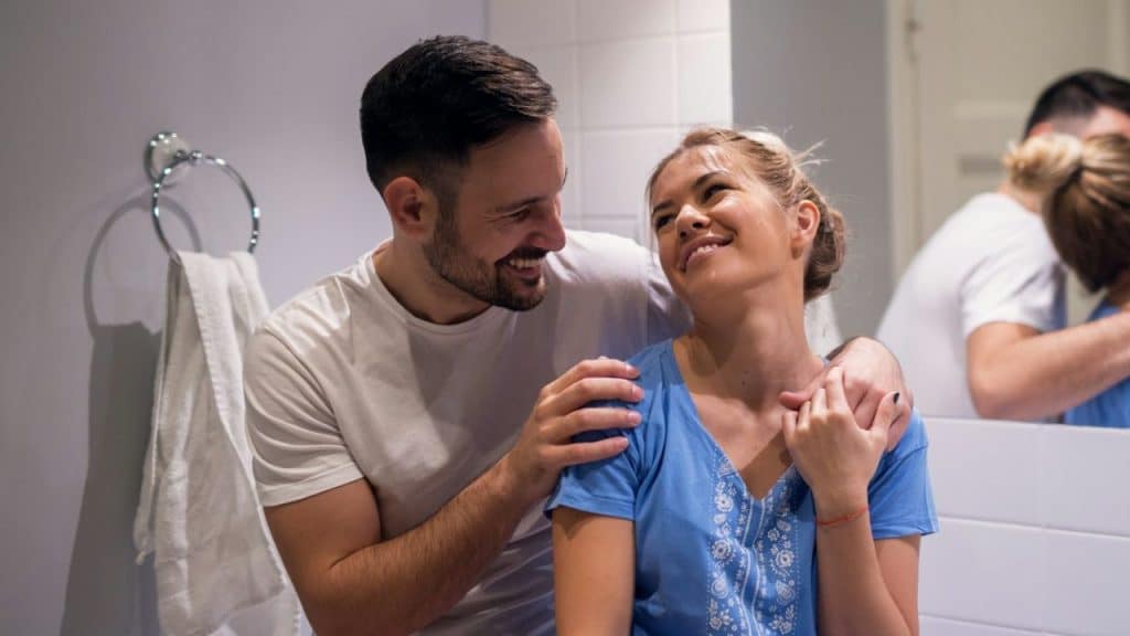 A smiling couple embraces in a bathroom, looking happily into each other's eyes.