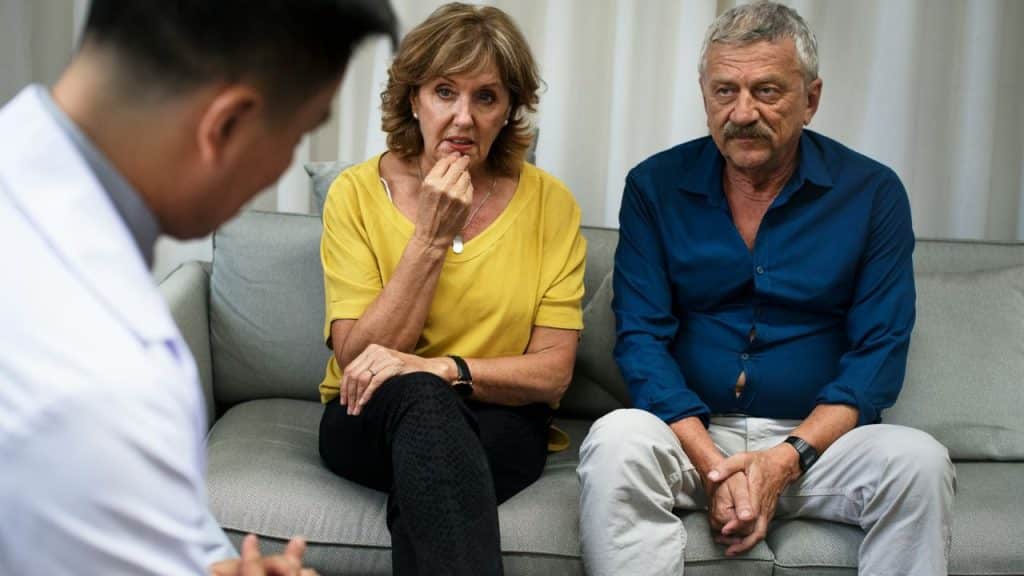 A mature couple sits on a couch, listening intently to a professional.