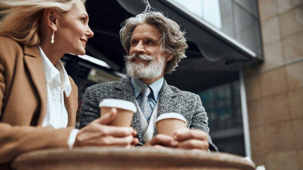 An older, stylishly dressed couple with coffee cups talks outdoors at a table.