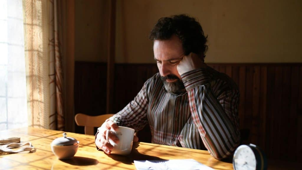 A middle-aged, bearded man sits alone at a table, looking distressed.