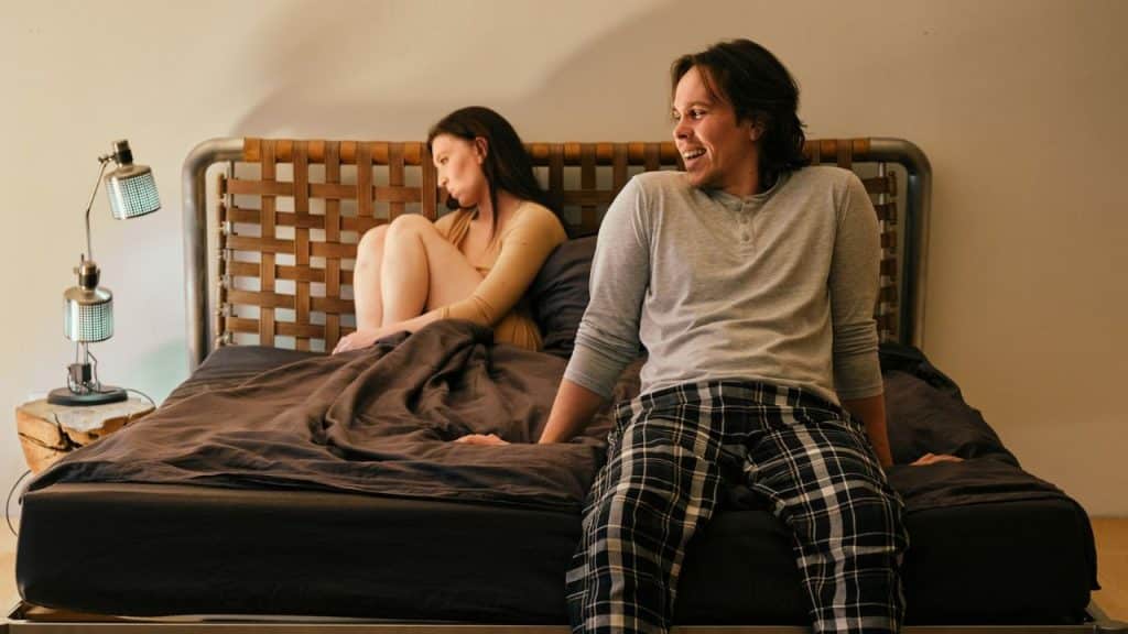 A smiling man in pajamas sits on a bed, looking back at a woman sitting with her back to him.
