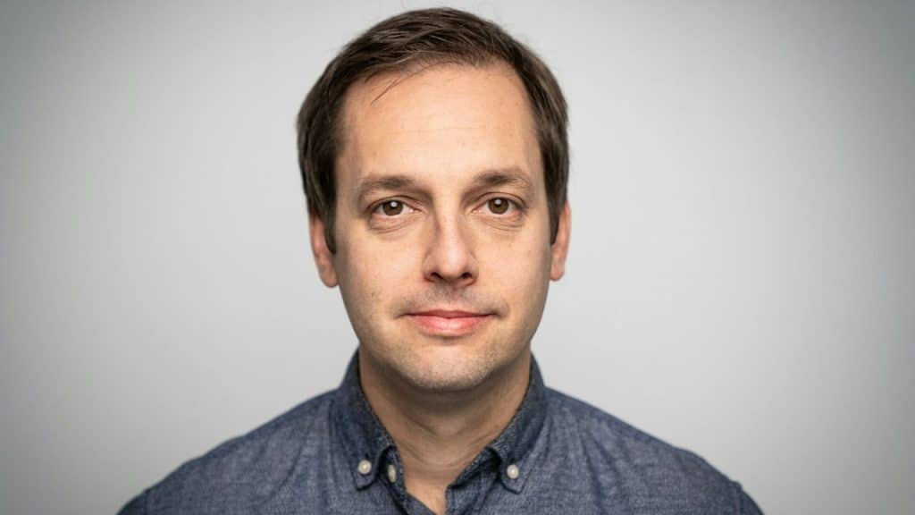 A headshot of a man with brown hair and a blue button-down shirt.