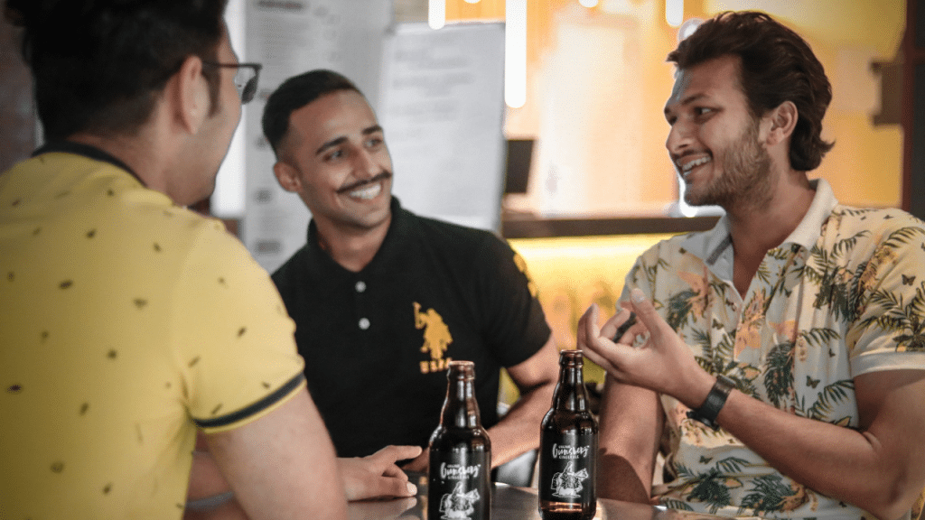 Three men sit around a table with beer bottles, smiling and talking.