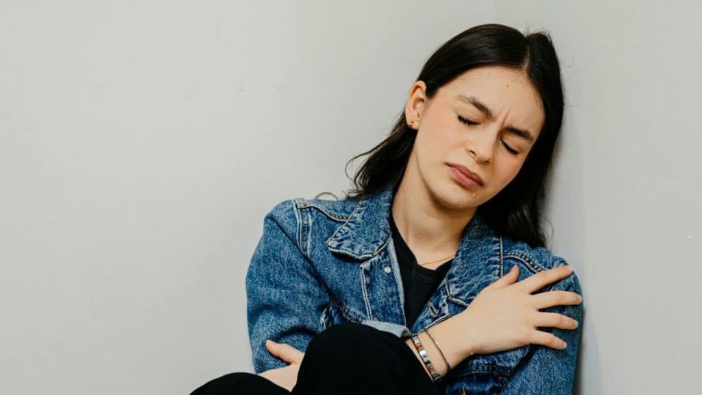 A young woman with a jean jacket is sitting on the floor in a corner, crying with her eyes closed.