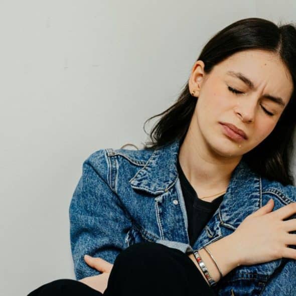A young woman with a jean jacket is sitting on the floor in a corner, crying with her eyes closed.
