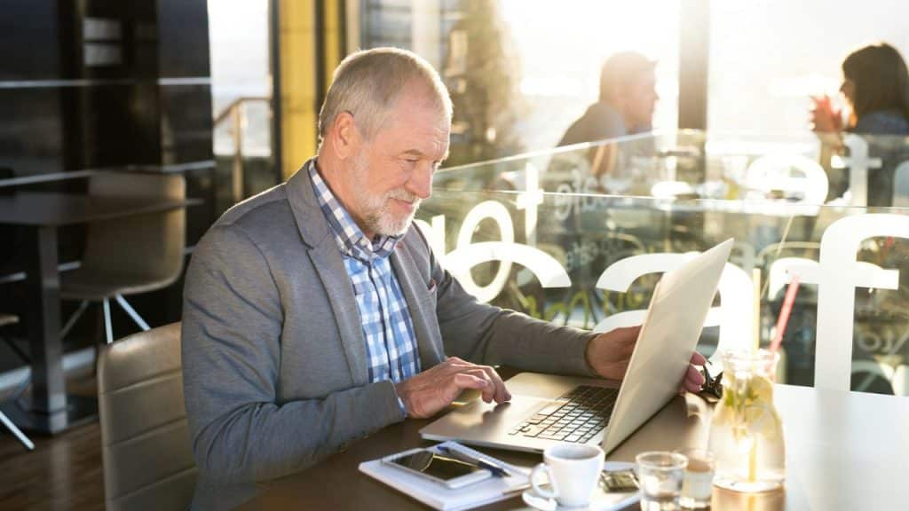A happy, senior businessman is working on his laptop at a sunny cafe.