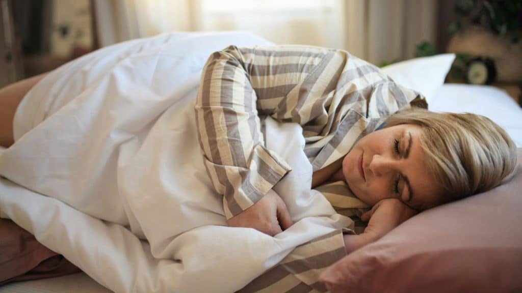 A woman with light hair is sleeping in a striped shirt and is covered by a white comforter. Her hands are near her face.