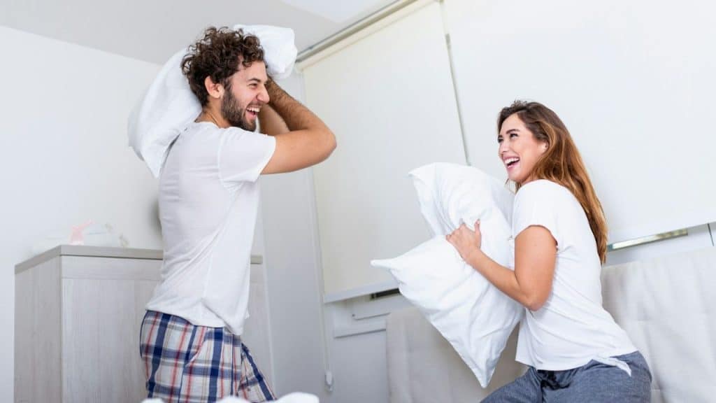 A smiling couple is having a pillow fight on a bed in a bright white room.