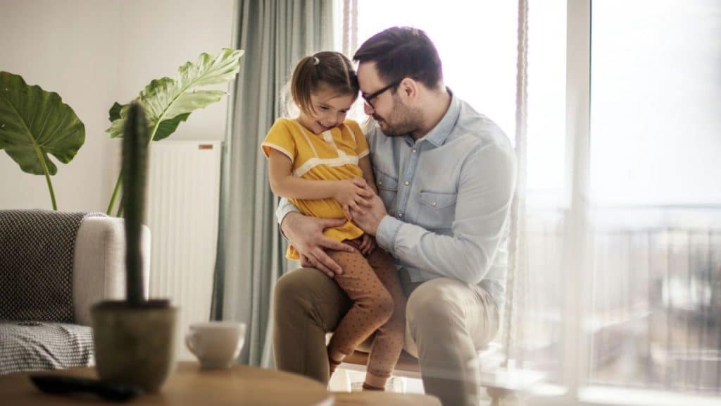 A smiling man sits indoors holding a young girl on his lap near a large window.