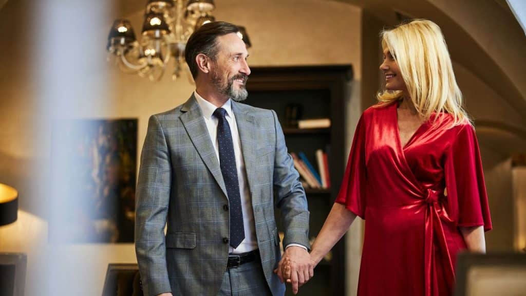 A smiling, elegantly dressed couple holds hands and looks at each other indoors.