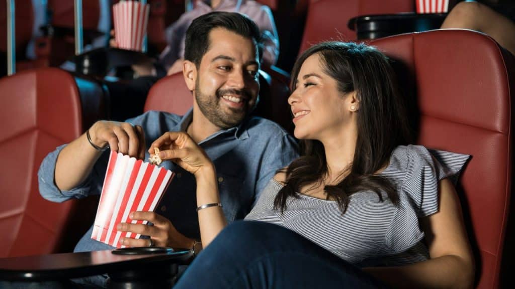 A smiling couple sits in a movie theater, sharing a bucket of popcorn.