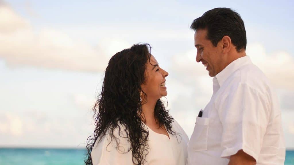 A joyful Hispanic couple smiles at each other by the turquoise ocean.