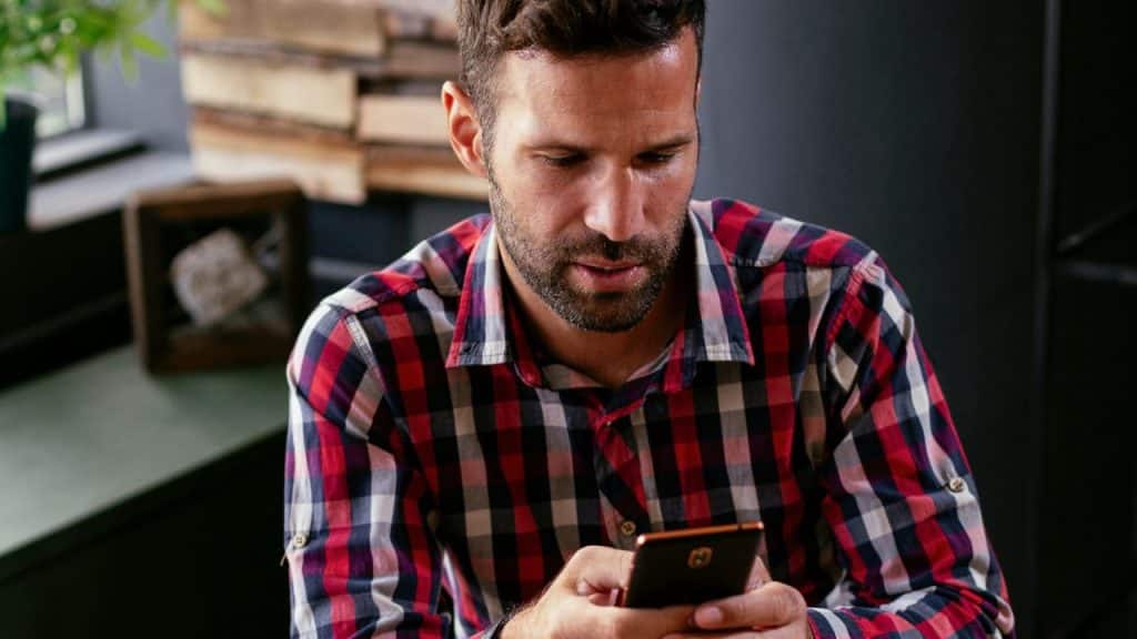 A focused man with a beard and plaid shirt looks down at a mobile phone in his hands.