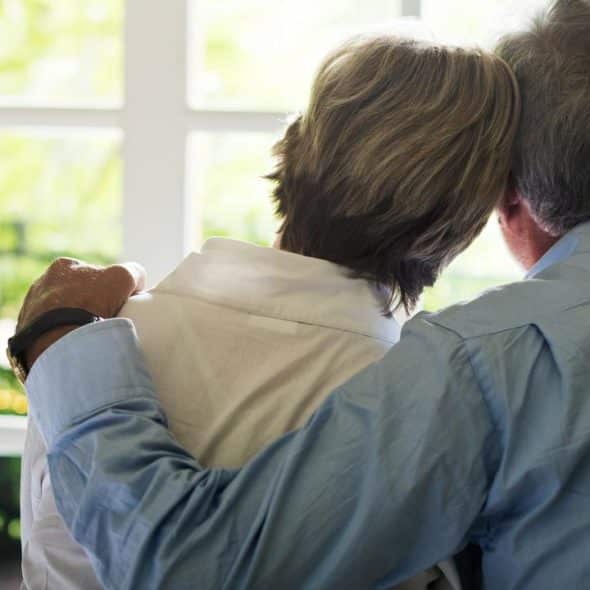 A senior couple sitting together by a window, with the man’s arm around the woman.