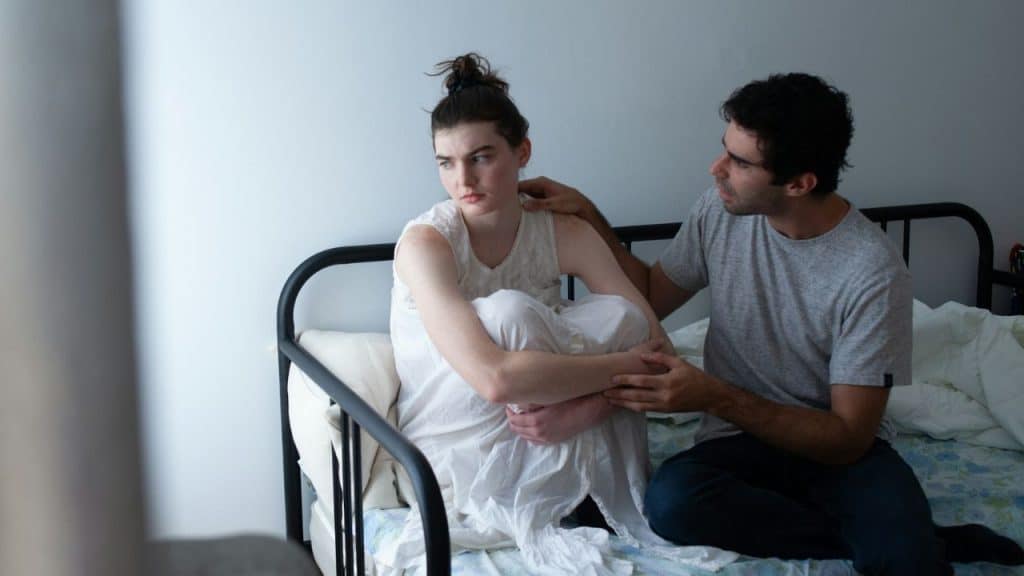 A sad-looking woman sits with her arms crossed while a man tries to comfort her.