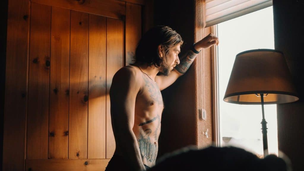 A shirtless man with tattoos stands inside, leaning on a window frame and looking out.