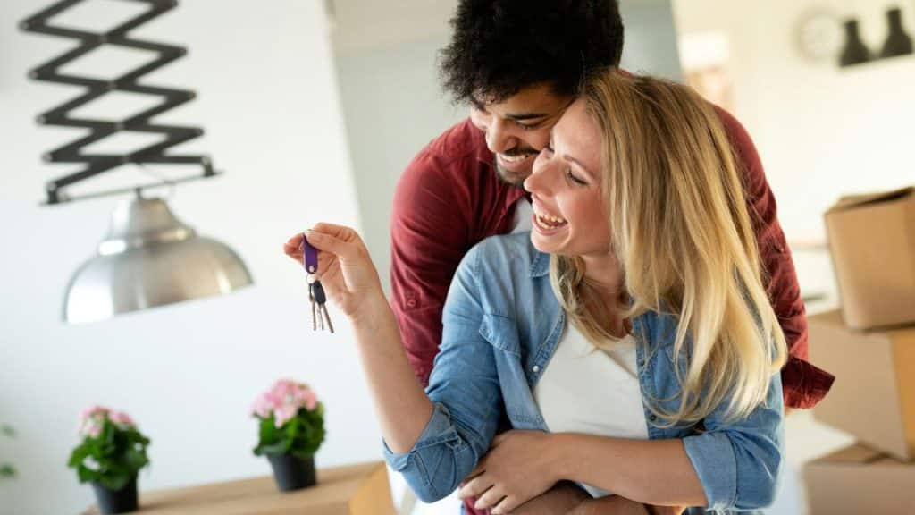 A happy couple smiles, and the woman holds up a set of keys. There are cardboard boxes behind them.