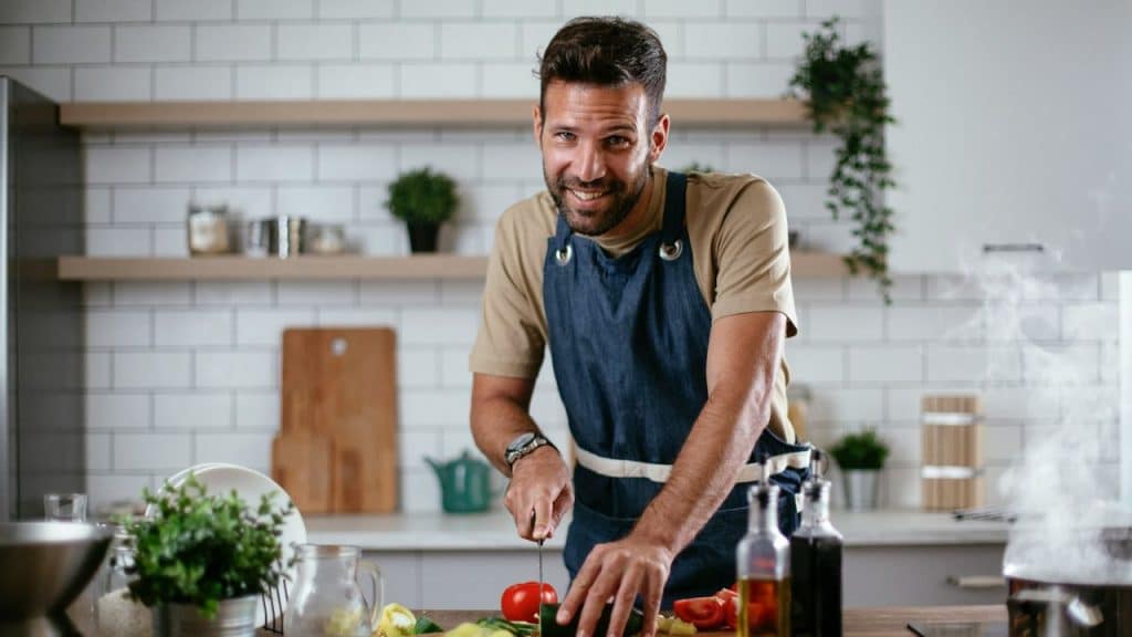 A man in a denim apron smiles at the camera while cutting a tomato in a kitchen.