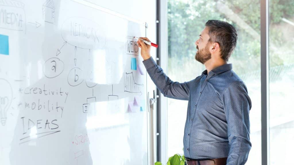 A businessman with a beard is writing on a whiteboard in an office.