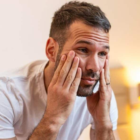 A man is looking in a mirror with his hands on his face, while a woman sits in the background.