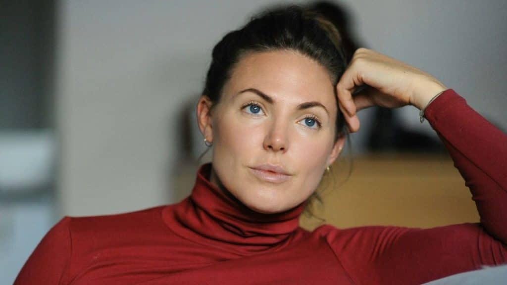 A woman in a red turtleneck rests her head on her hand, looking off to the side.