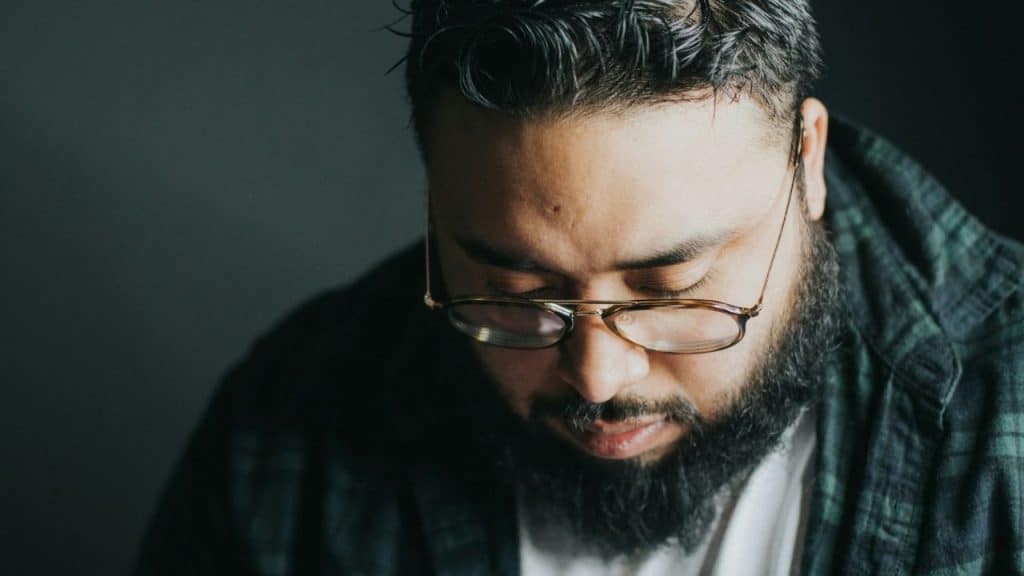 A man with a full beard and glasses looks down in a close-up indoor shot.