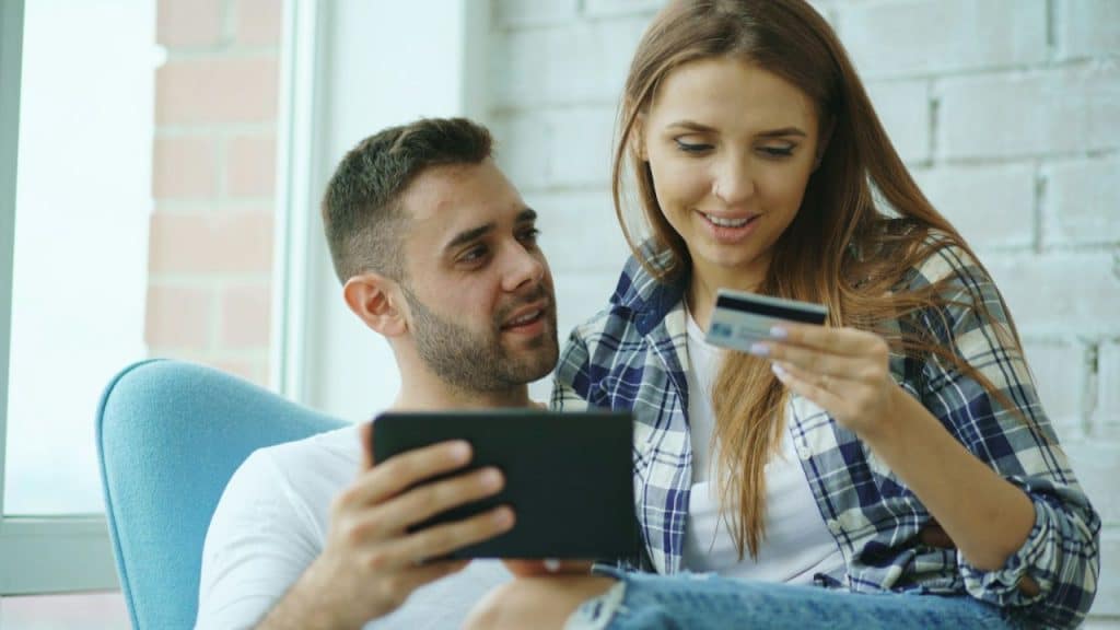 A smiling couple sits together, looking at a tablet and holding a credit card.