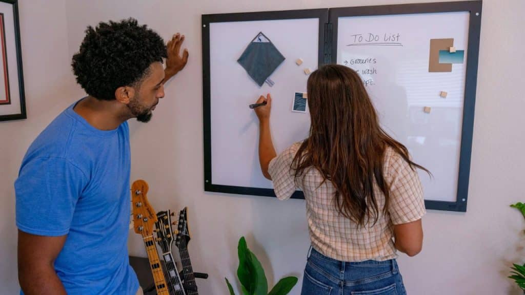 A man watches as a woman writes on a To Do List whiteboard.