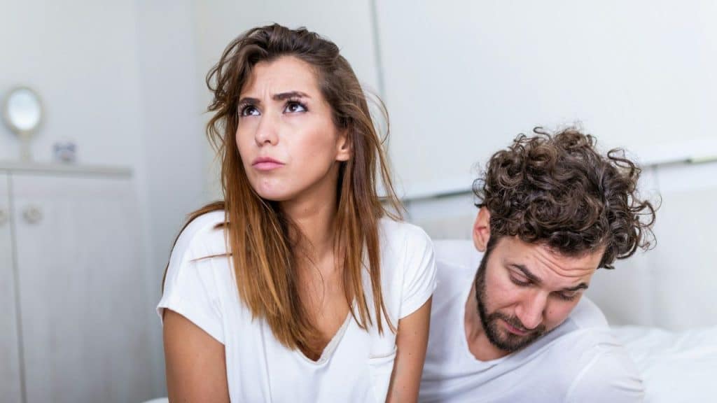 A young woman looks up with a disgusted expression while a man lies on the bed behind her.