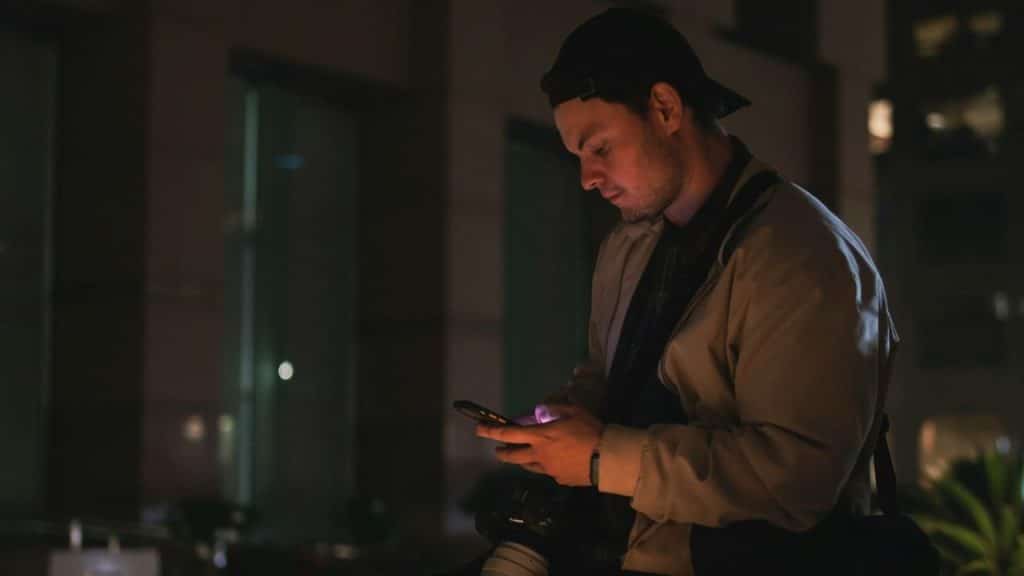 A man wearing a baseball cap looks down at his phone at night.