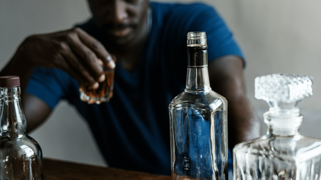 A blurred man holds a glass of whiskey, sitting in front of three bottles of alcohol.