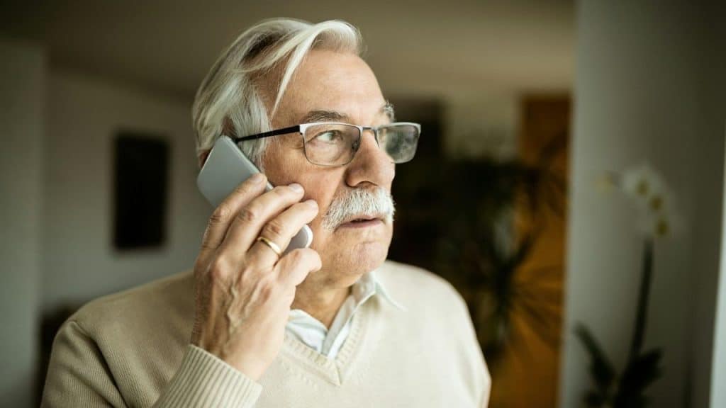 A thoughtful senior man with a mustache and glasses holds a phone to his ear.