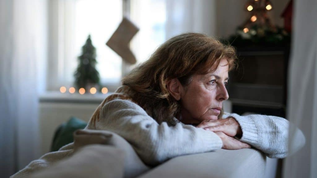 A woman with long, brown hair sits on a couch, resting her head on her hands with a solemn expression.
