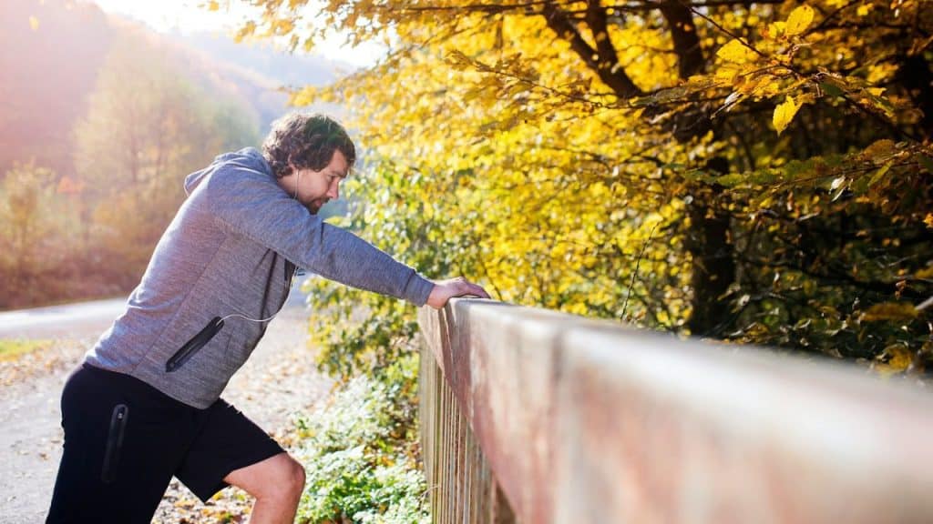 A man in athletic clothes stretches against a railing on a country road in autumn.
