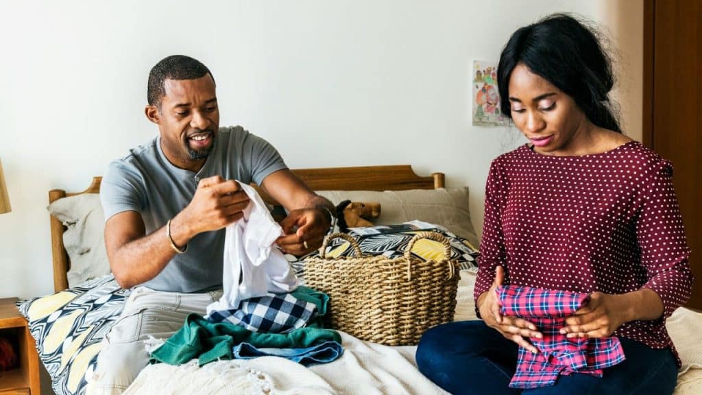 A man and a woman are sitting on a bed, folding laundry from a wicker basket.