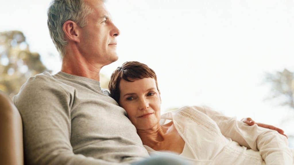 A smiling, middle-aged woman is resting her head on a man's shoulder as they sit together.