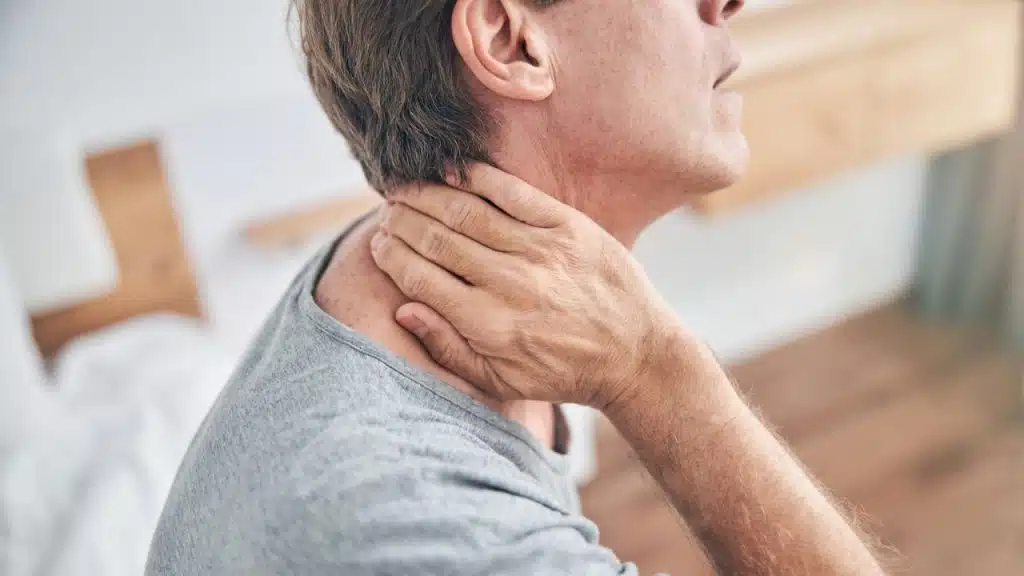 A man in a gray shirt touches his neck, appearing to be in pain.