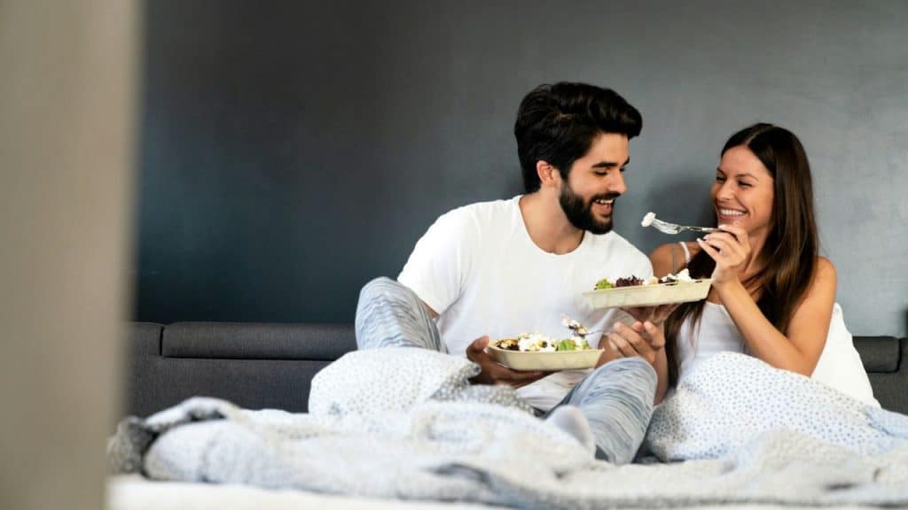 A happy couple sits in bed and eats salad. The woman is holding a forkful of food up to the man.