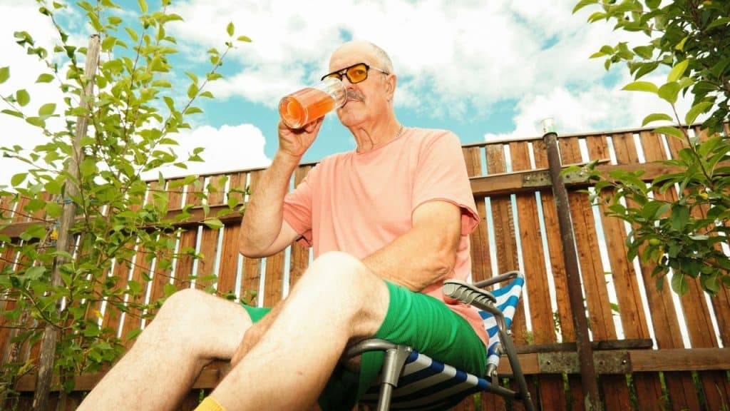 A bald man with a mustache and sunglasses drinks from a glass while sitting on a chair outdoors.