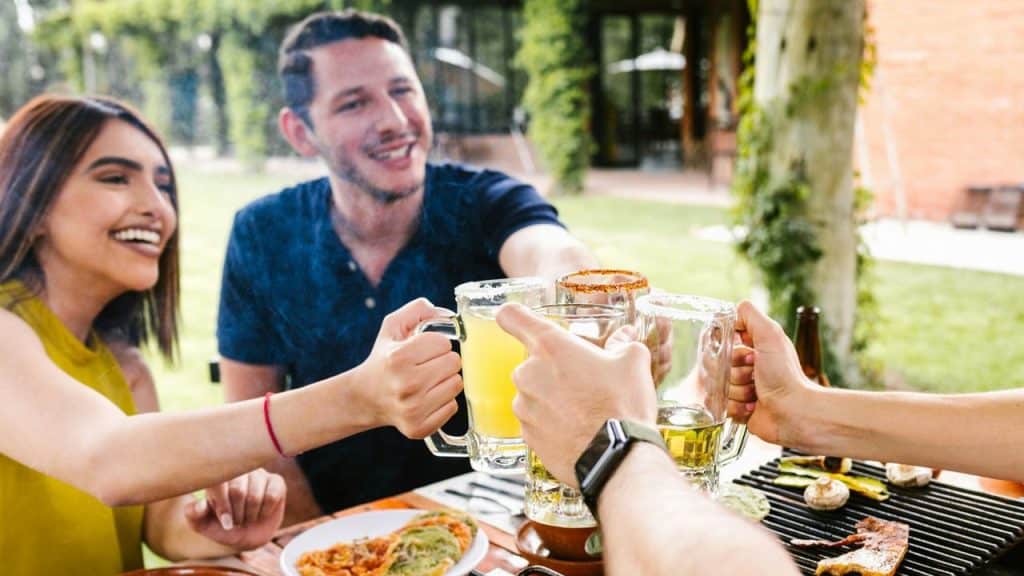 A group of people sits at a picnic table, laughing and toasting with drinks.
