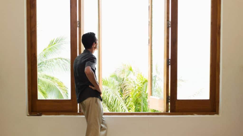 A man stands at an open window, looking out at the palm trees.