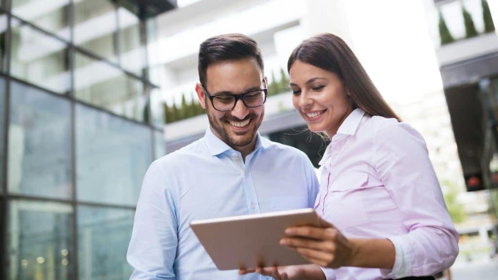A smiling man and woman stand together, looking at a tablet computer.