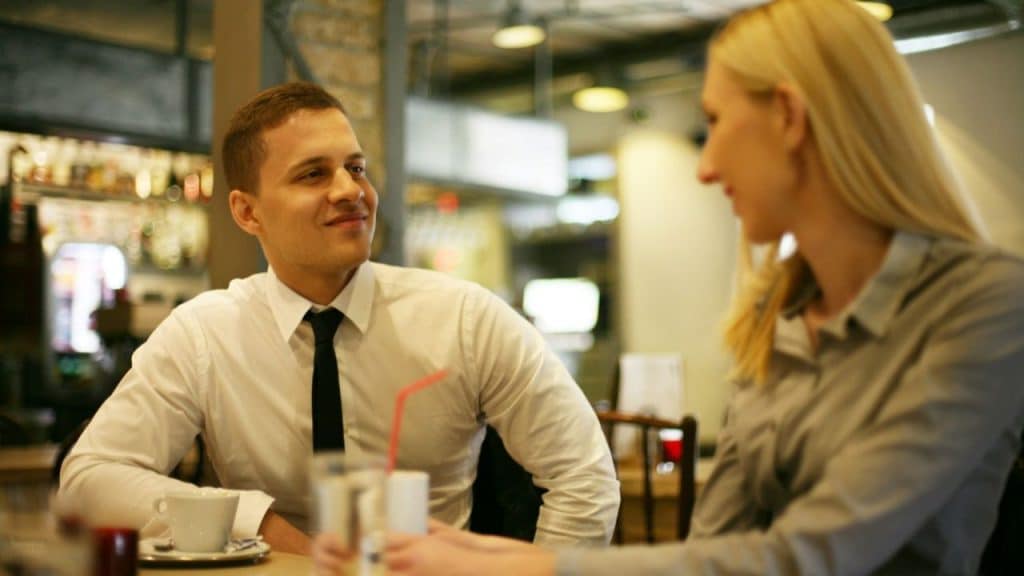 A man in a white shirt and tie smiles at a woman across a table at a cafe.