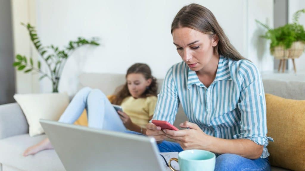A woman on her phone and a girl on a tablet sit on a couch.