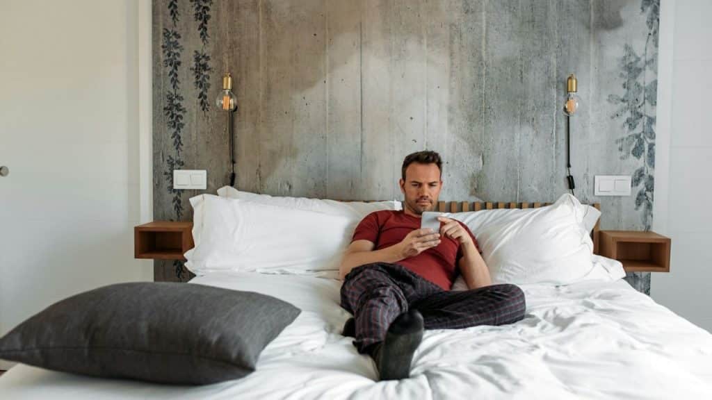A man in pajamas is sitting on a bed and looking at his phone.